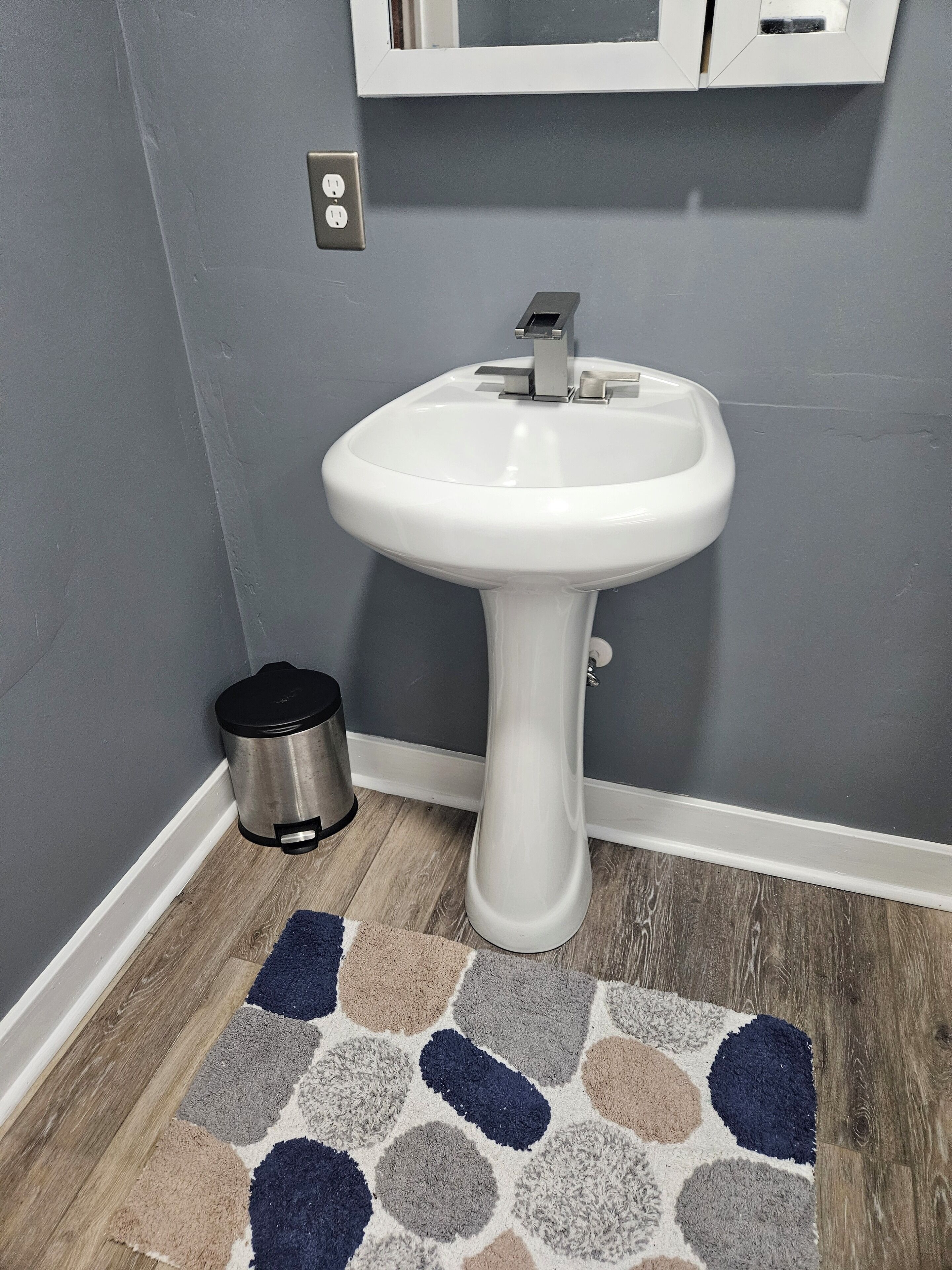 A bathroom with a white pedestal sink, a trash can, and a patterned rug on the floor.