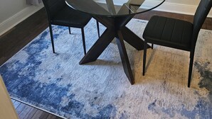 Een eethoek met een ronde glazen tafel, vier zwarte stoelen, een blauw tapijt met patroon en een kunstwerk in een lijst aan de muur.
