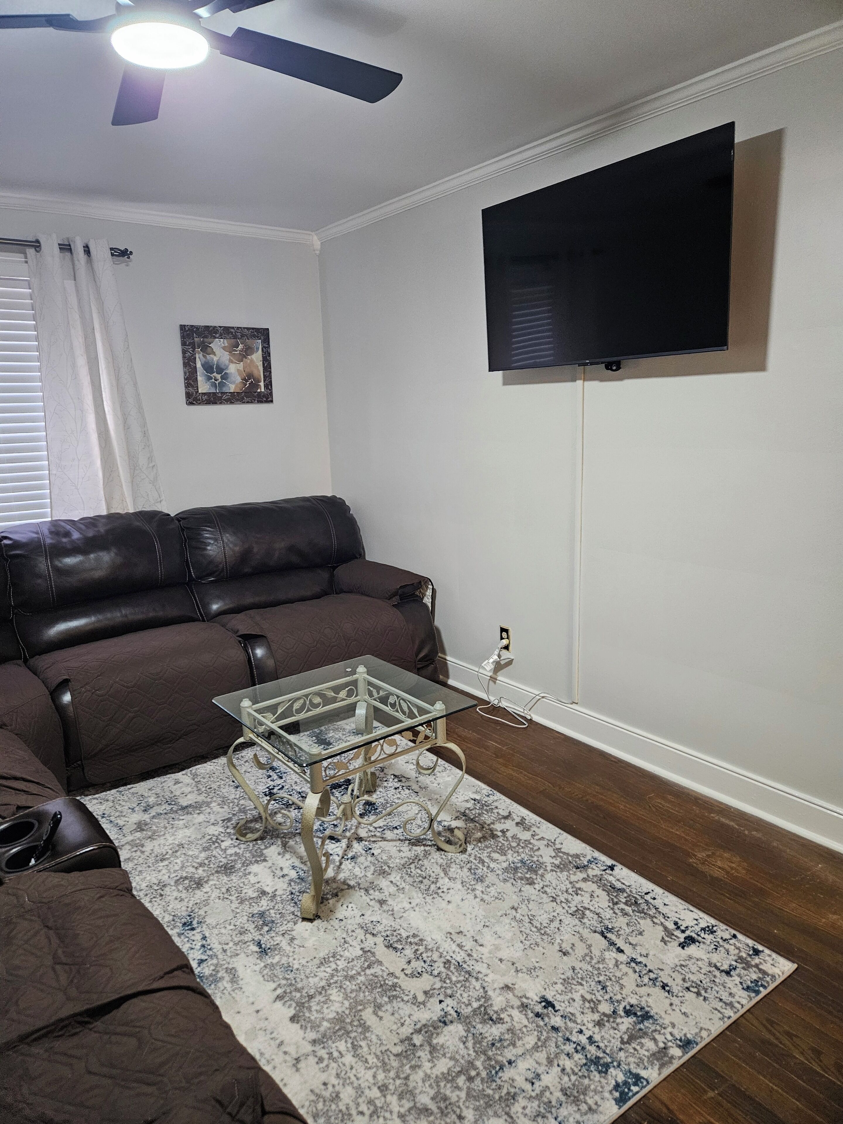 A living room with a brown leather sofa, a glass coffee table, a patterned rug, a ceiling fan, a flat-screen TV mounted on the wall, and a window with white curtains.