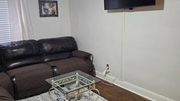 A living room with a brown leather sofa, a glass coffee table, a patterned rug, a ceiling fan, a flat-screen TV mounted on the wall, and a window with white curtains.