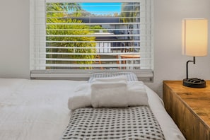 A hotel room with a bed, a window with blinds, a bedside table with a lamp, and a view of greenery outside. - Modern 2BR in South Tampa with Pool, Parking & Workspace (Tampa)