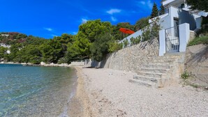 Un bâtiment situé sur la côte, doté d’un escalier en pierre menant à un balcon qui offre une vue sur une plage de galets et sur des eaux d’un bleu limpide.