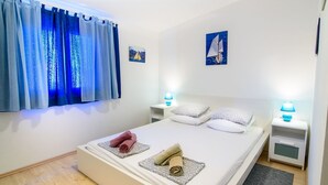 Una cama bien hecha con almohadas estampadas y una toalla enrollada, una mesita de noche con lámpara, y una ventana con cortinas azules.
