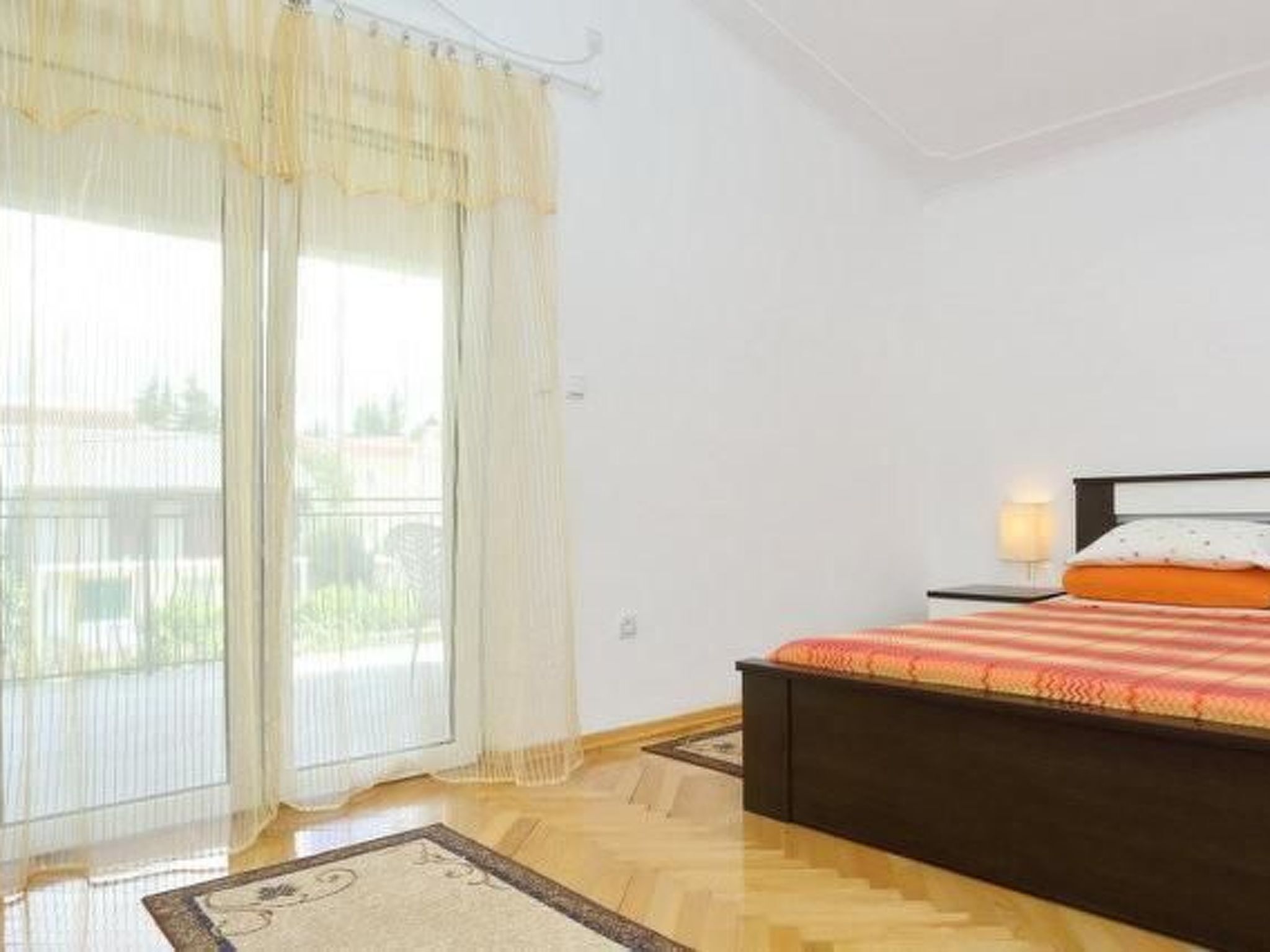 Ein Schlafzimmer mit einem Bett, einem Nachttisch, einer Lampe und Blick durch ein Fenster mit Vorhängen.