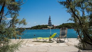 Una playa con dos sillas de descanso, una sombrilla a rayas y vistas al mar y árboles lejanos.