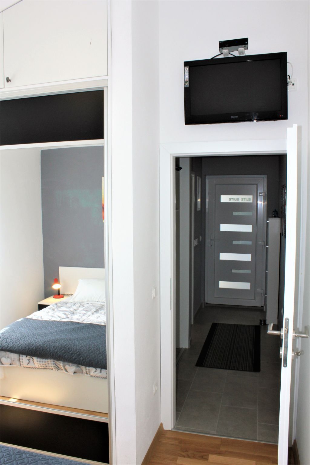 A bedroom with a bed, a bedside table with a lamp, and a TV mounted above the door.