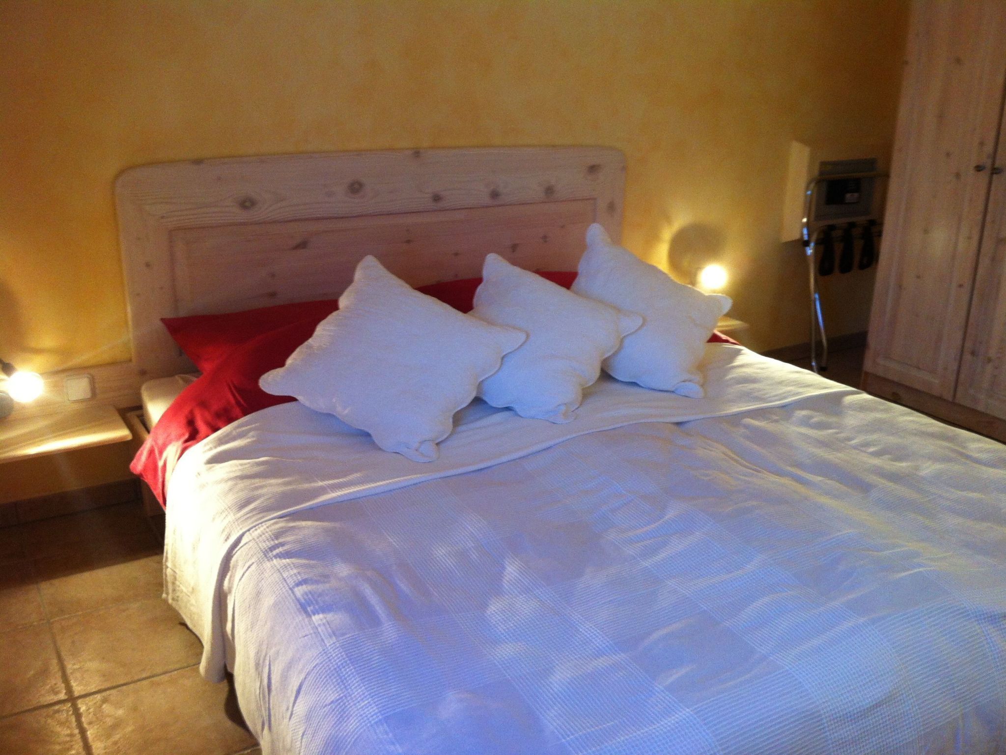 A neatly made bed with white bedding and red pillows, a wooden headboard, and a bedside table with a lamp.