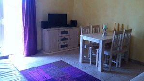 Un coin repas avec une table et des chaises en bois, un téléviseur posé sur un meuble, et un rideau violet.