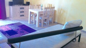 Un lit simple avec une tête de lit, une petite table de chevet et une chaise. On trouve également une table à manger avec quatre chaises et un téléviseur posé sur un meuble.