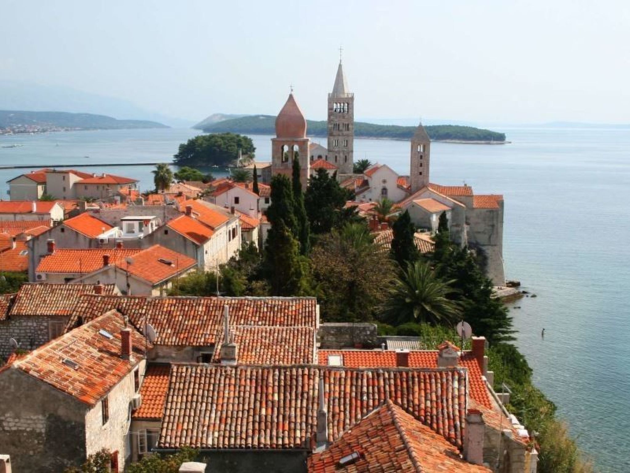Une ville côtière aux bâtiments aux toits de tuiles rouges, une église imposante dotée d’un clocher et une mer calme en toile de fond.