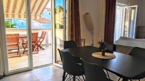 Un coin repas avec une table et des chaises, une vue à travers une porte-fenêtre coulissante donnant sur un espace extérieur aménagé avec des chaises et une table en bois, et une grande fenêtre avec des rideaux.