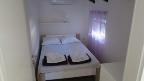 En liten, välordnad säng med vita sängkläder och en svart sänggavel, ett fönster med lila gardiner och en väggmonterad luftkonditioneringsanläggning.