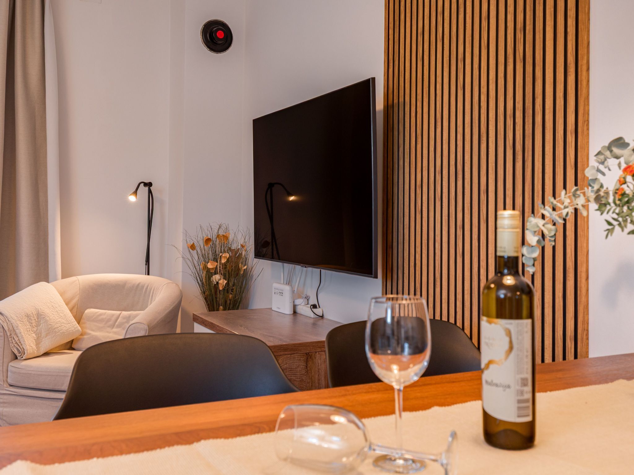 A modern living room with a flat-screen TV, a wooden panel wall, a sofa, a coffee table, and a wine bottle.