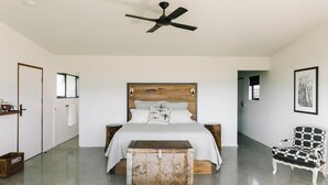 Un dormitorio con una cama grande, cabecera de madera, cómoda, alfombra blanca, sillón negro y blanco, y ventilador de techo.