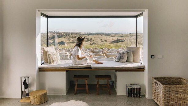 Una habitación con un ventanal grande que da a un paisaje rural, un banco de madera y un taburete pequeño.