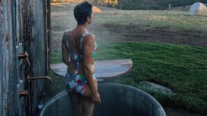 Una persona en traje de baño parada en un jacuzzi con una ducha sobre ella, con vista a un campo cubierto de césped y colinas al fondo.