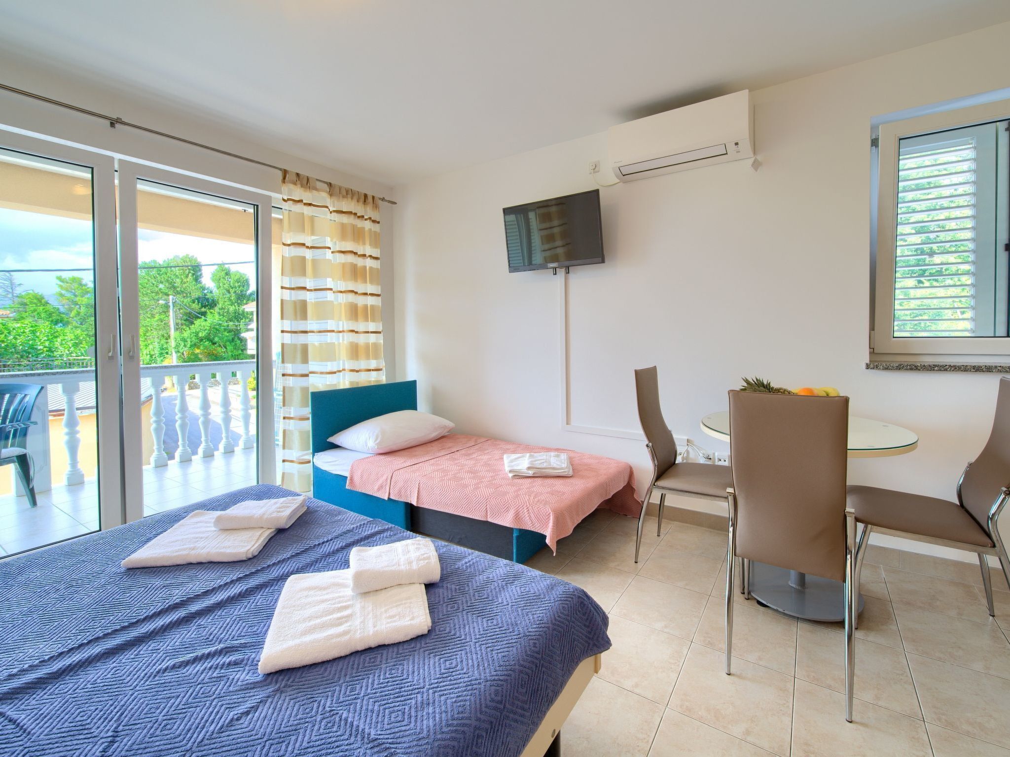 Habitació d'hotel amb balcó, un llit amb capçalera blava, una taula petita amb cadires i un televisor fixat a la paret.