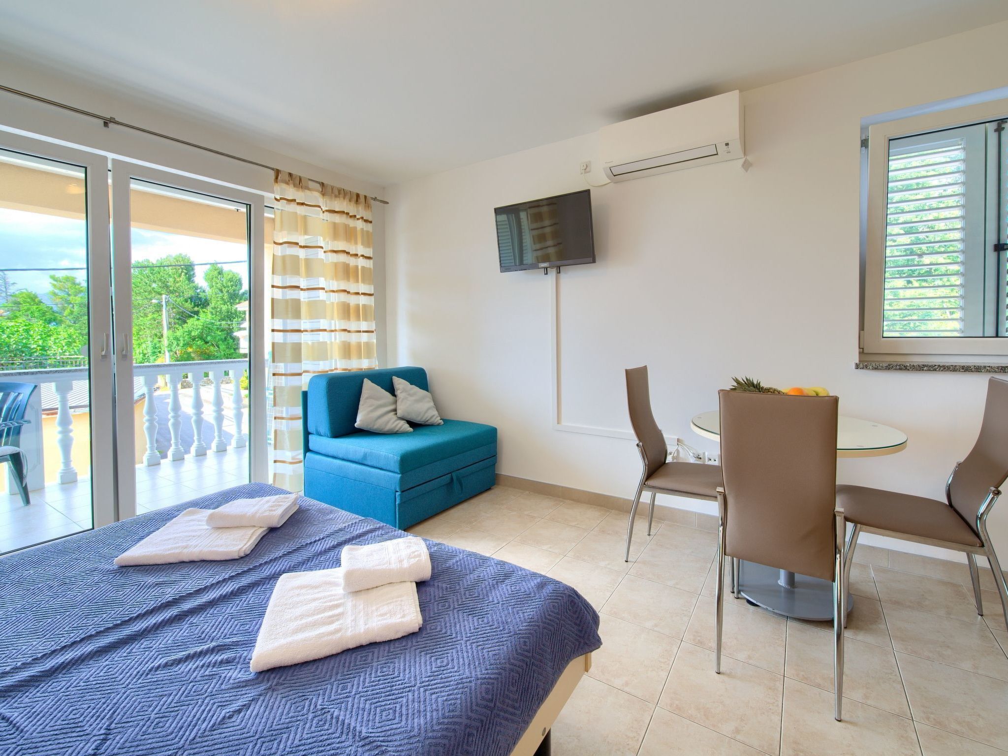 Une chambre d’hôtel moderne, équipée d’un lit, d’un canapé bleu, de deux fauteuils, d’une petite table et d’un balcon avec vue.