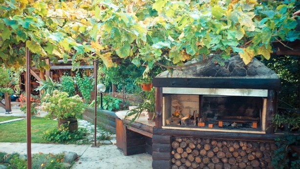 A rustic outdoor seating area with a stone fireplace, surrounded by a garden with various plants and a wooden pergola overhead.