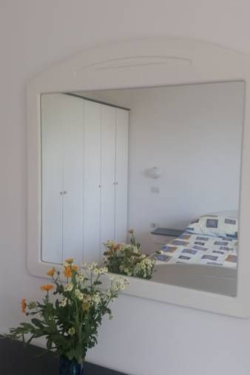 Une chambre à coucher avec un grand miroir, un lit recouvert d’une couvre-lit à motifs et un vase rempli de fleurs posé sur une commode.