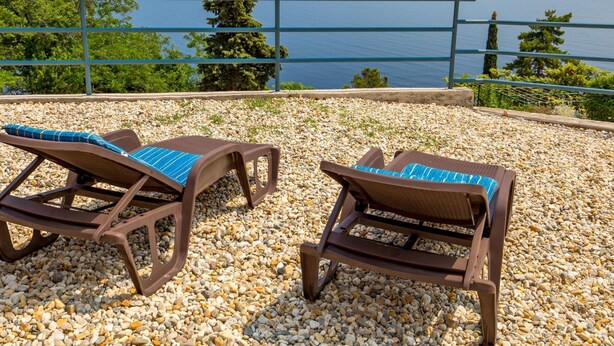 To komfortable loungestole med blå hynder på en stenbelagt terrasse med udsigt over vand og fjerne landskaber.