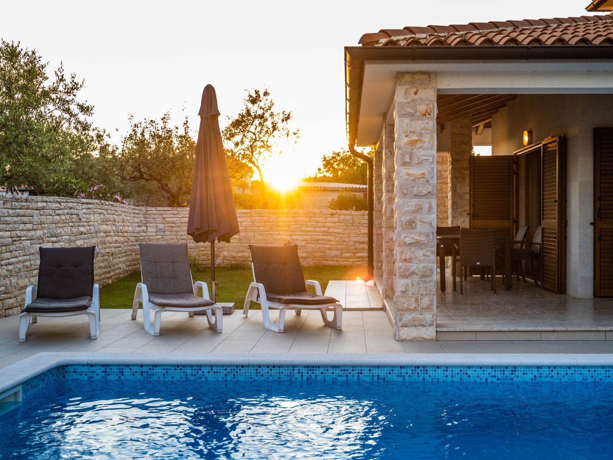 A poolside area with lounge chairs and an umbrella, a stone wall, and a building with a tiled roof.