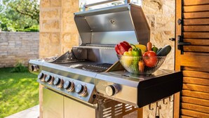 Ein Gasgrill im Freien mit einer Gemüsekörbchen-Auflage, vor einer Steinsäule und einer Holztür.