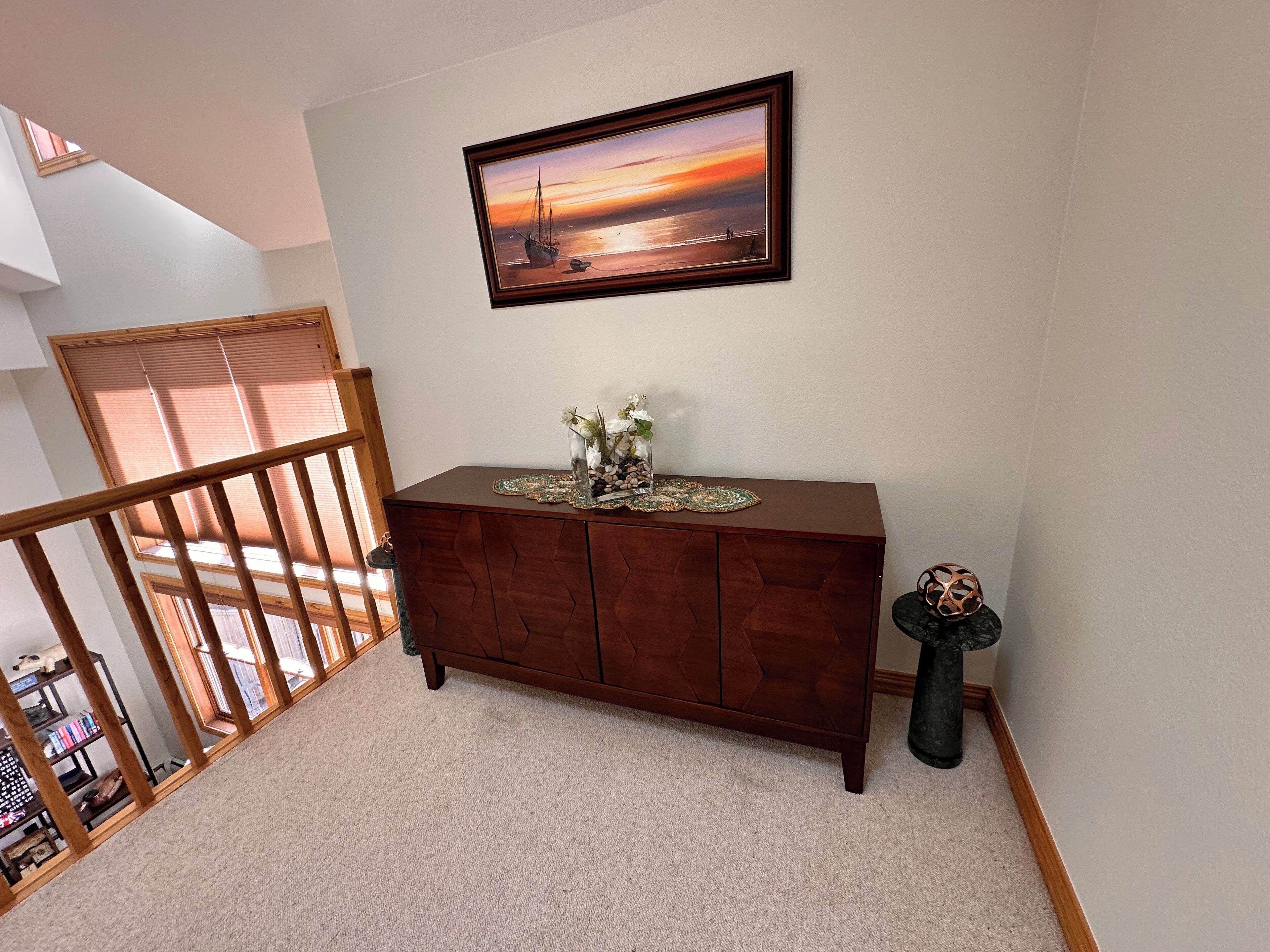 A wooden console table with a vase of flowers, a framed painting of a sunset, and a small decorative item on a carpeted floor.