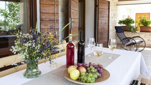 Una mesa dispuesta con botellas de vino, copas y una bandeja de fruta, con contraventanas de madera y una pared de piedra al fondo.
