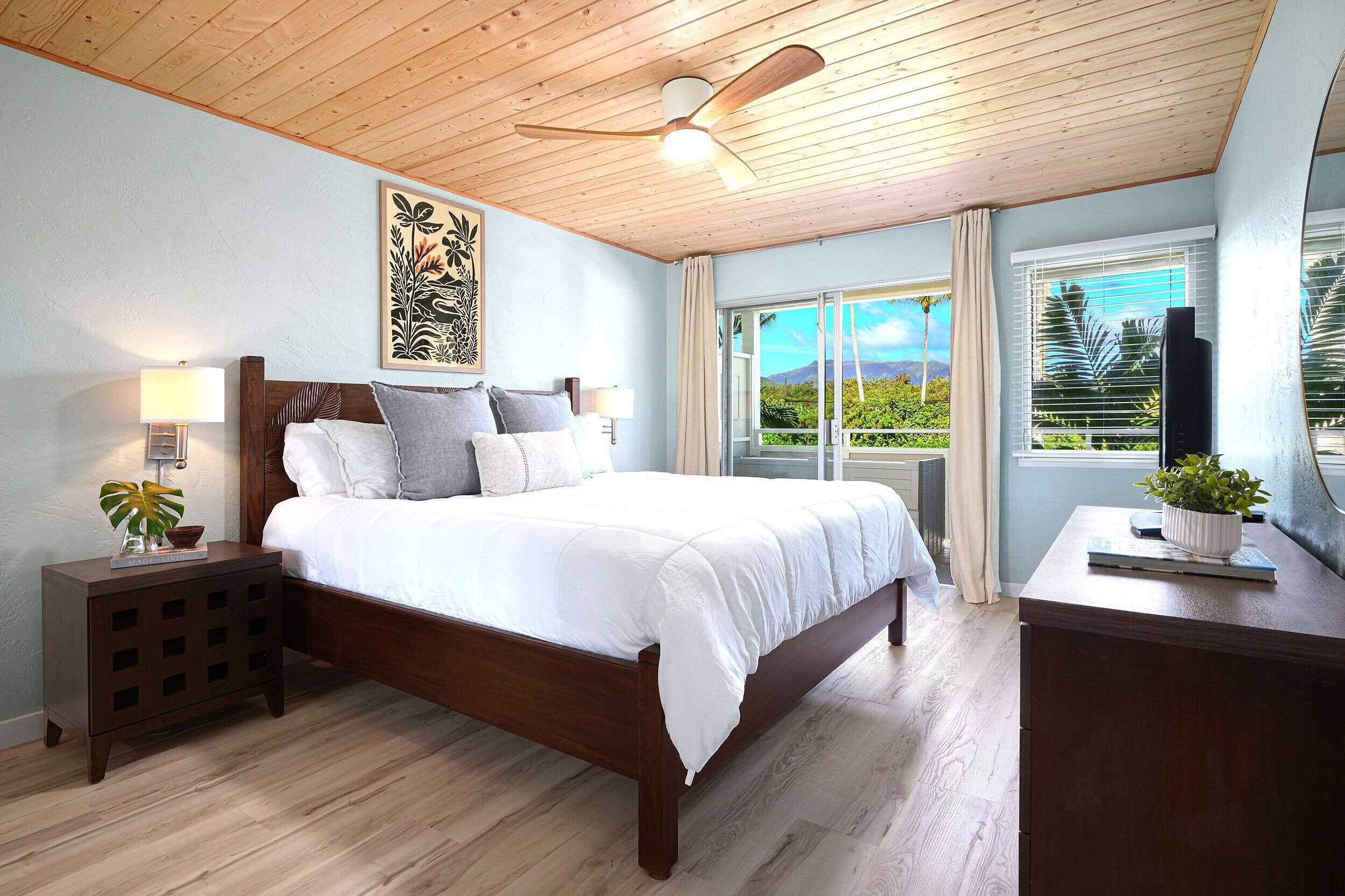 A bedroom with a wooden ceiling fan, a bed with white bedding, a nightstand with a lamp, and a view of greenery through the window.