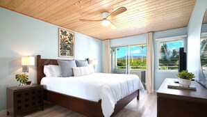 Une chambre avec un ventilateur de plafond en bois, un lit avec une literie blanche, une table de chevet avec une lampe, et une vue sur la verdure par la fenêtre.