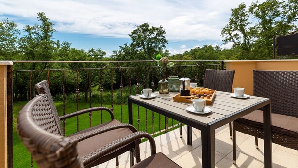 A terrace with a table set for breakfast, wicker chairs, and a view of a lush garden. - Modernes Apartment mit Balkon und Gemeinschaftsgarten am Waldrand by Interhome (Dracevac)