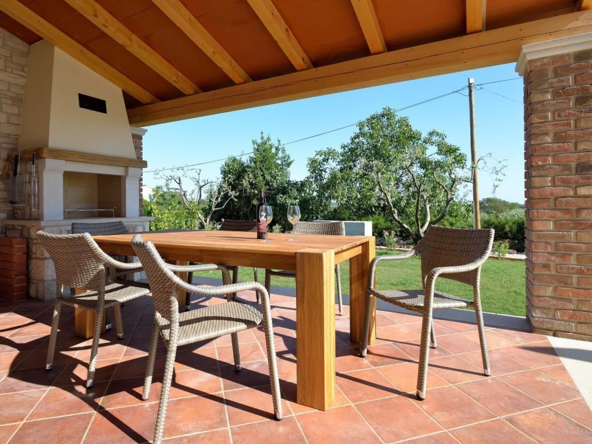 Een terras buiten met een houten tafel en rieten stoelen, een barbecue en uitzicht op bomen en een heldere lucht.