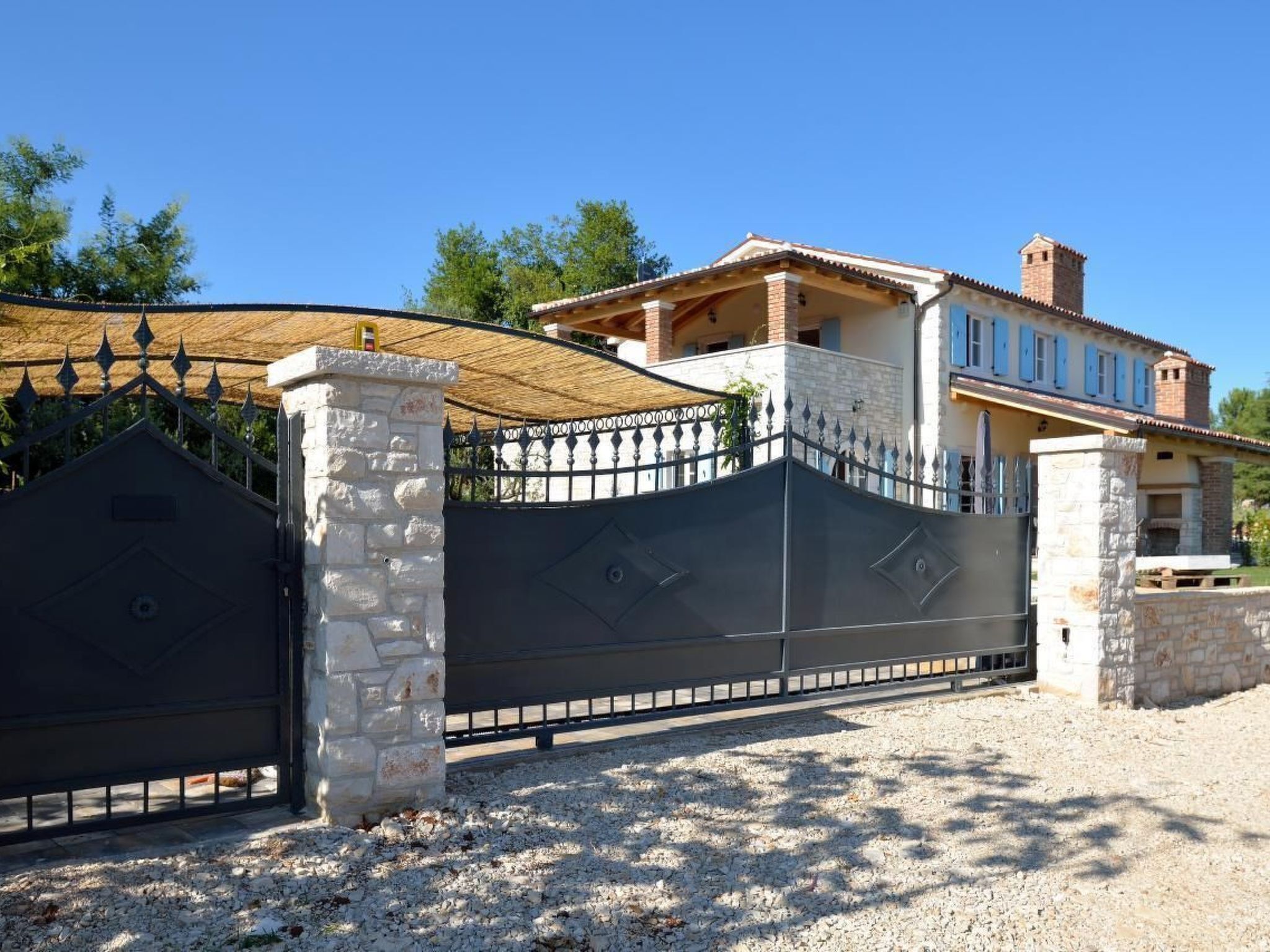 A gated property with a stone pillar, a metal gate, and a building with a tiled roof and a chimney.