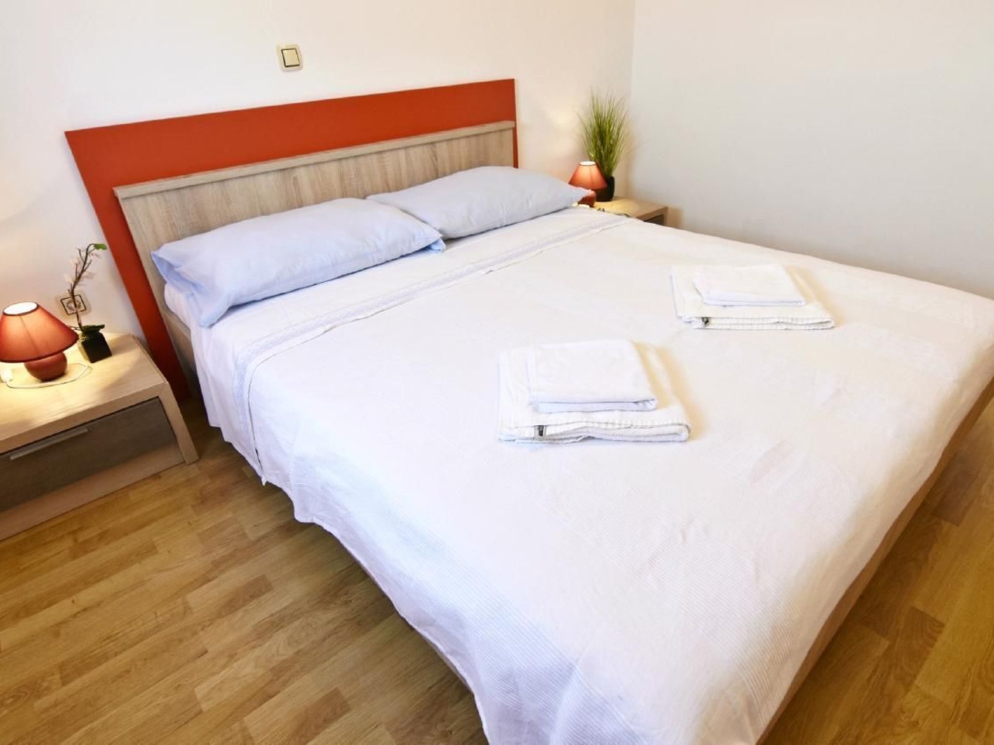 A neatly made bed with white linens and two folded towels, a red headboard, a bedside lamp, and a small plant on the nightstand.