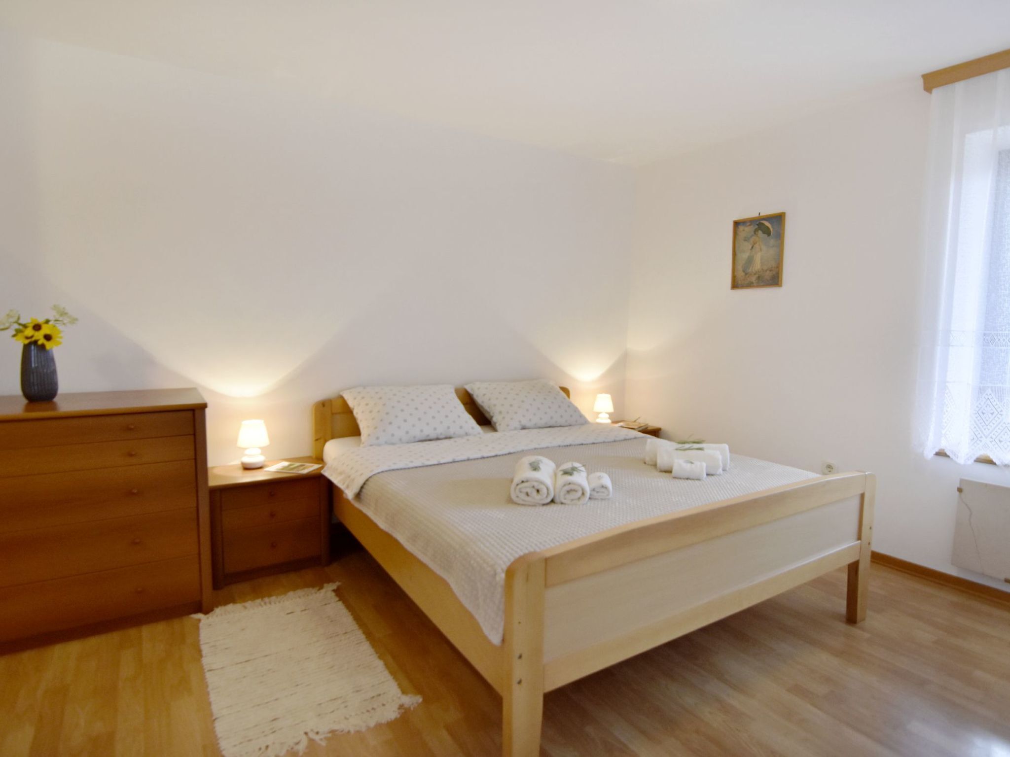 Une chambre à coucher avec un lit en bois, une table de chevet, une commode, un vase de fleurs et une fenêtre ornée de rideaux légers.