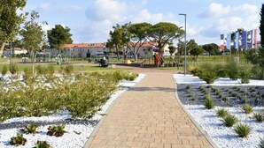 Um caminho pavimentado leva a um parquinho infantil com um escorrega vermelho e balanços, cercado por áreas ajardinadas e edifícios ao fundo.