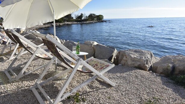 Una spiaggia con un ombrellone bianco, poltroncine di legno e un tavolino con una bottiglia e un bicchiere.