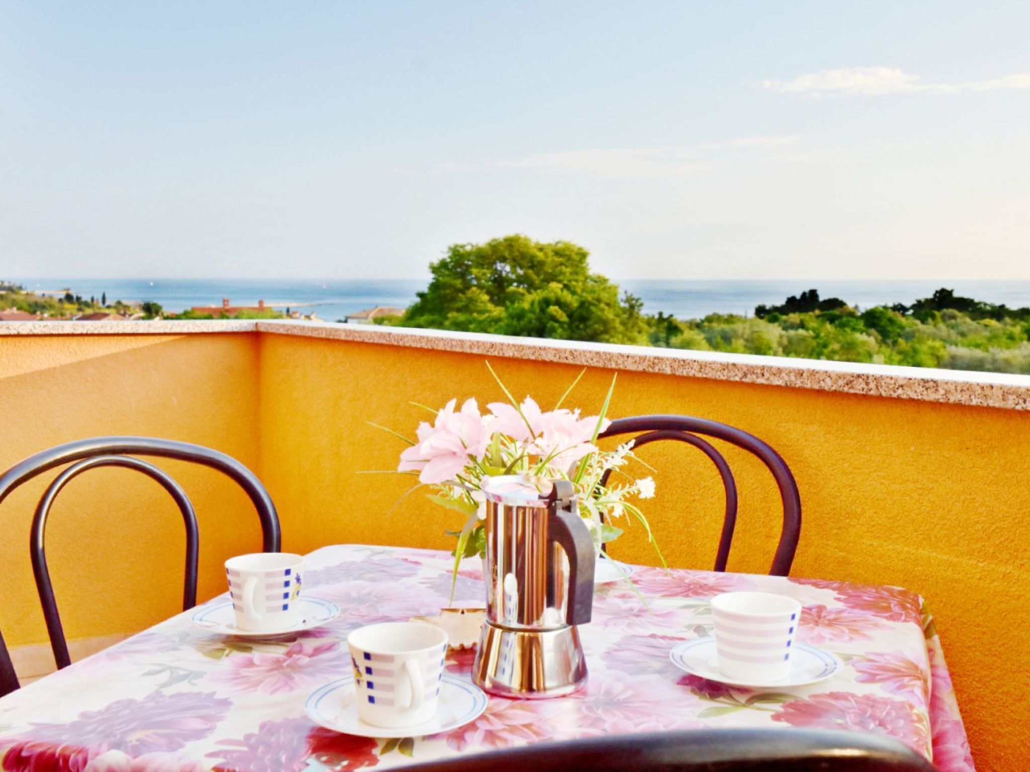 Une table dressée pour deux, recouverte d’une nappe à motifs floraux, agrémentée d’un vase rempli de fleurs et d’une carafe, le tout placé sur un balcon offrant une vue panoramique.