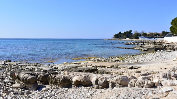 A rocky beach with a clear blue sea, a boat, and a building with trees. - Appartement in Zambratija mit Schöner Terrasse by Interhome (Umag/Zambratija)