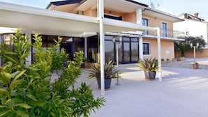 Ein zweistöckiges Haus mit überdachter Terrasse, Kübelpflanzen und blauem Himmel.