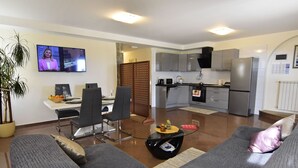 Ein modernes Wohnzimmer mit grauem Sofa, einem Essbereich mit Tisch und Stühlen, einer Küche mit grauen Schränken und einem Kühlschrank sowie einem an der Wand befestigten Fernseher.