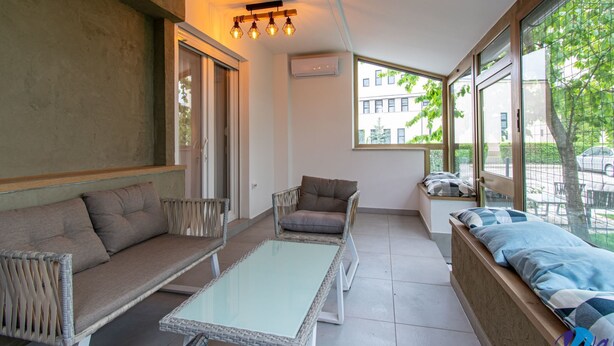 Una sala de estar moderna con sofá, sillón y una mesa de centro de vidrio. Amplias ventanas ofrecen vistas al exterior.