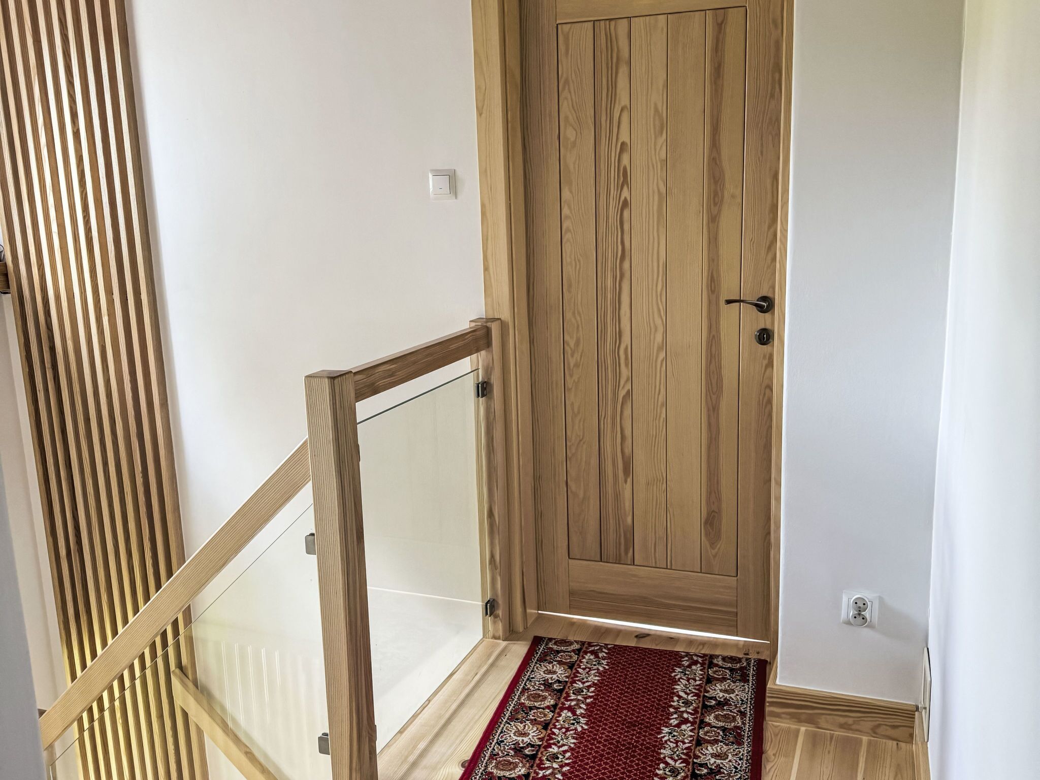 A wooden door with a glass panel, a wooden staircase with a glass railing, and a patterned rug on the floor.