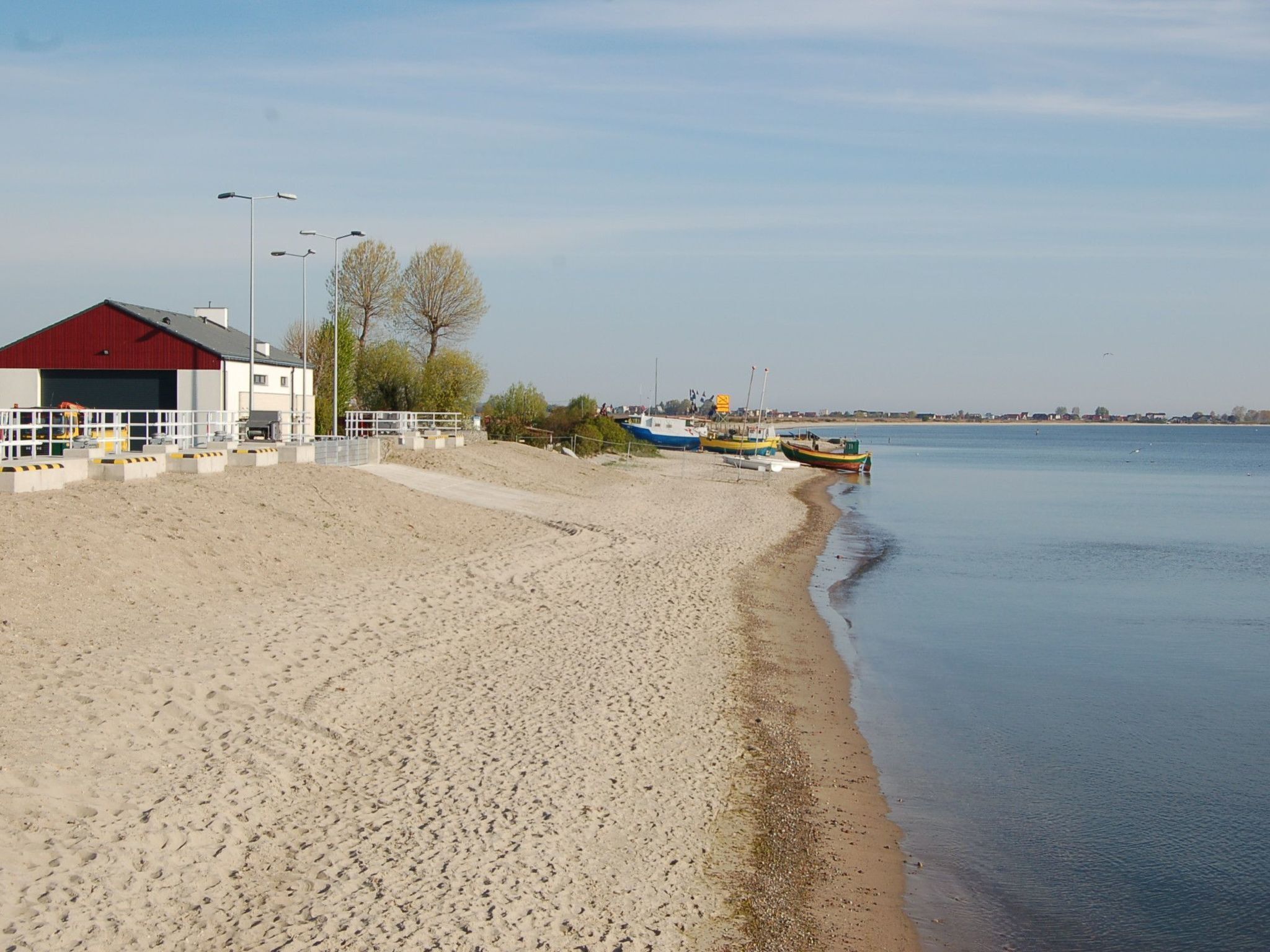 En sandstrand med en röd byggnad, en rad båtar och ett lugnt hav.