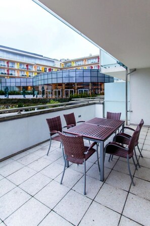 Un patio al aire libre con una mesa y sillas, baranda de vidrio y vista a un edificio de varios pisos con balcones coloridos.