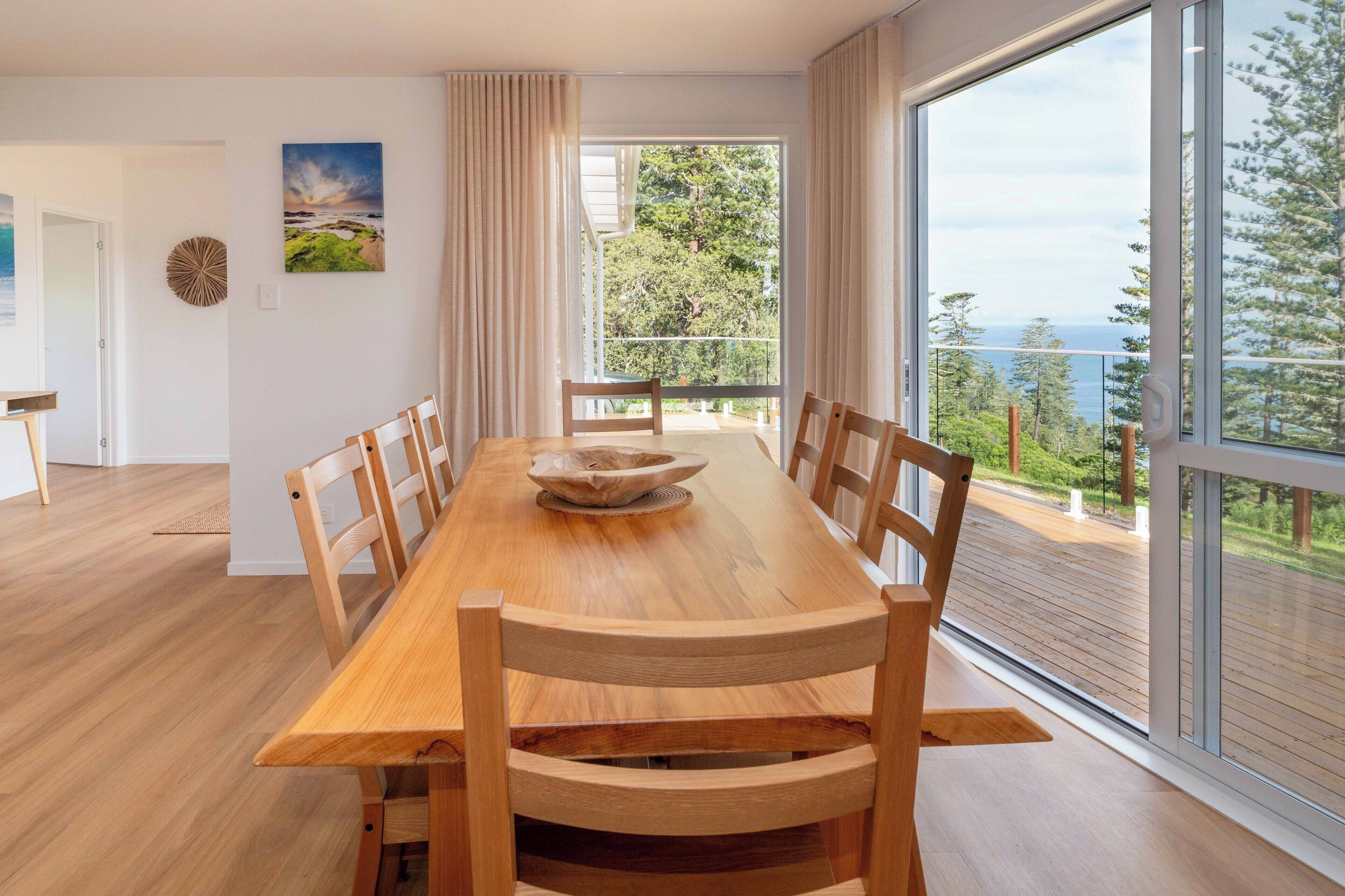 Ein Essbereich mit Holztisch und Stühlen, großen Fenstern mit Blick ins Freie und einem Bild an der Wand.