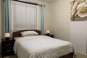 Une chambre à coucher comprenant un lit, deux tables de chevet, une fenêtre avec des stores, une lampe fixée au mur et une grande œuvre d’art à motifs floraux.