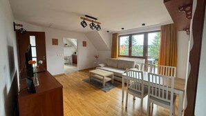 Un salon moderne avec un canapé, une table à manger et des chaises, un téléviseur, et une cuisine en arrière-plan.