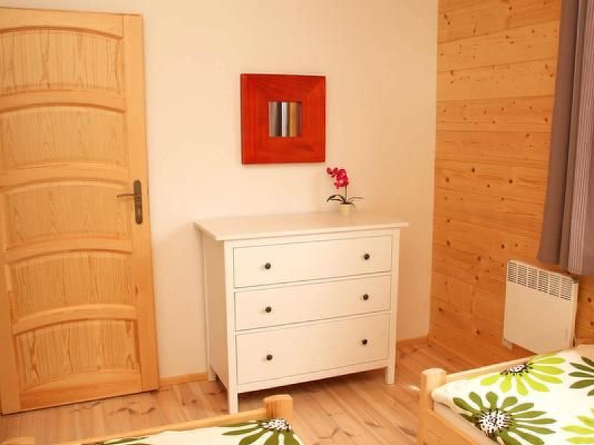 Ein Zimmer mit Holztür, einer weißen Kommode mit Schubladen, einem kleinen Bild in rotem Rahmen und einem Bett mit floralem Überwurf.